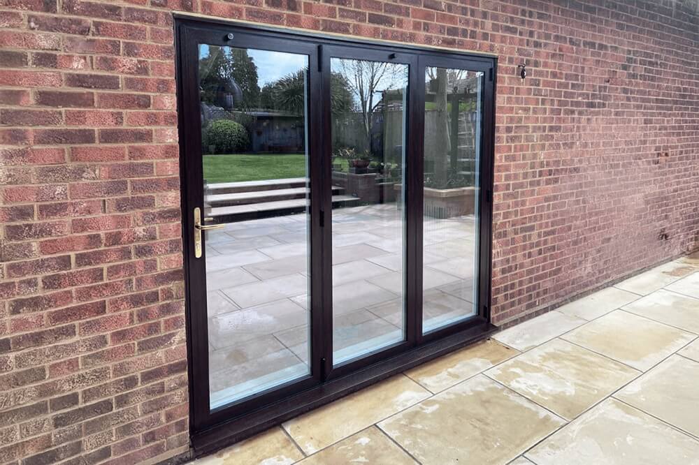Black aluminium bifold doors installed in a brick-built property, connecting an indoor space to a paved patio with a garden view.