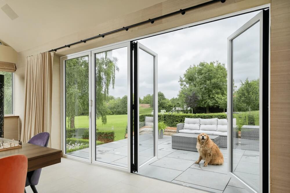 White sheerline bifold doors fully open, connecting a modern interior to an outdoor patio with a garden view.